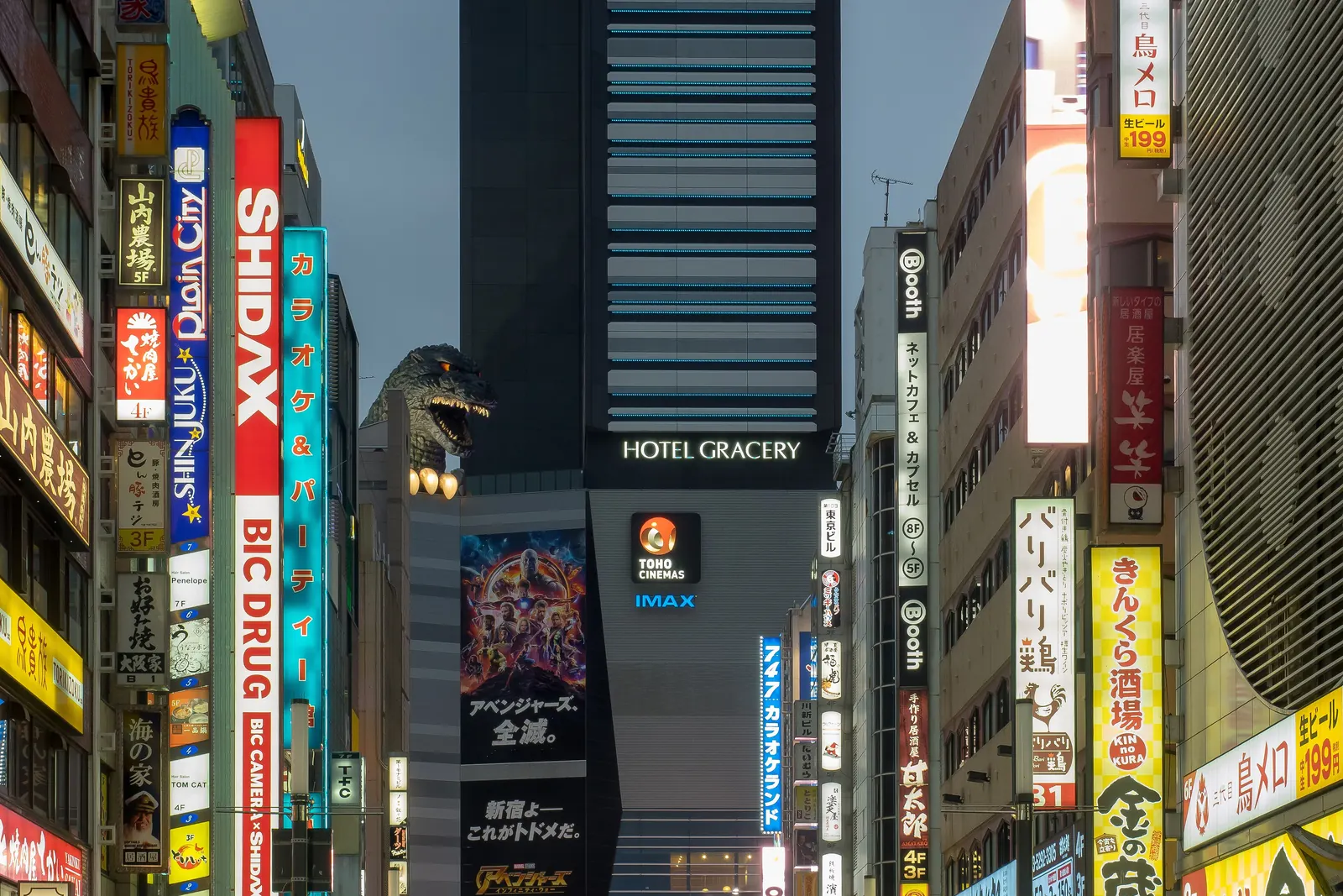 A Godzilla head looking over the rooftops of Shinjuku beside a hotel tower, above a street of vertical signs