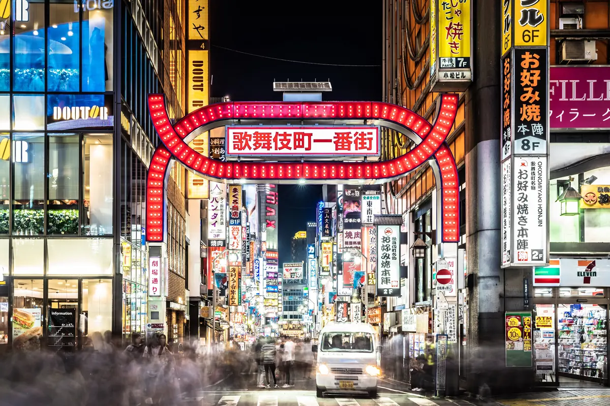 The red arched Kabukichō Ichibangai gate lit at night, with neon signs along the street beyond it and a white van waiting at the crossing