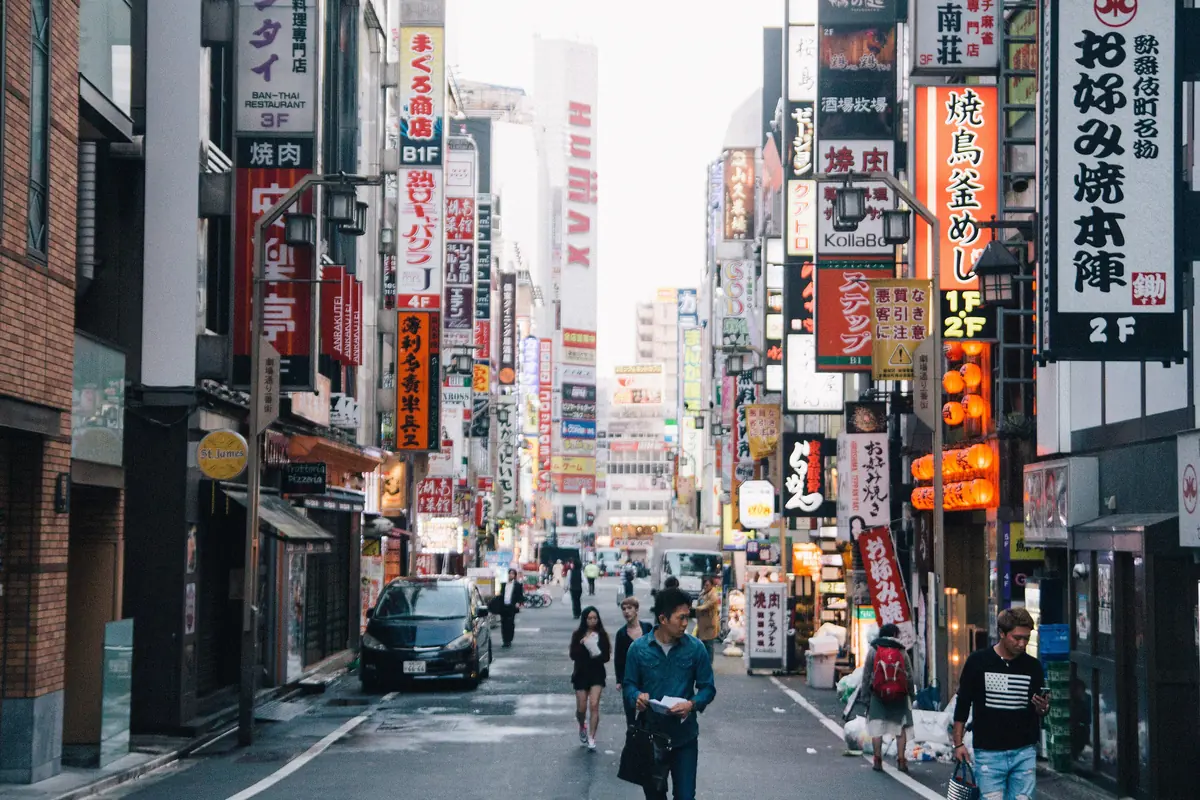 A quiet Kabukichō street in daylight, with cars parked along it, a few pedestrians and tall signs reading in Japanese on both sides