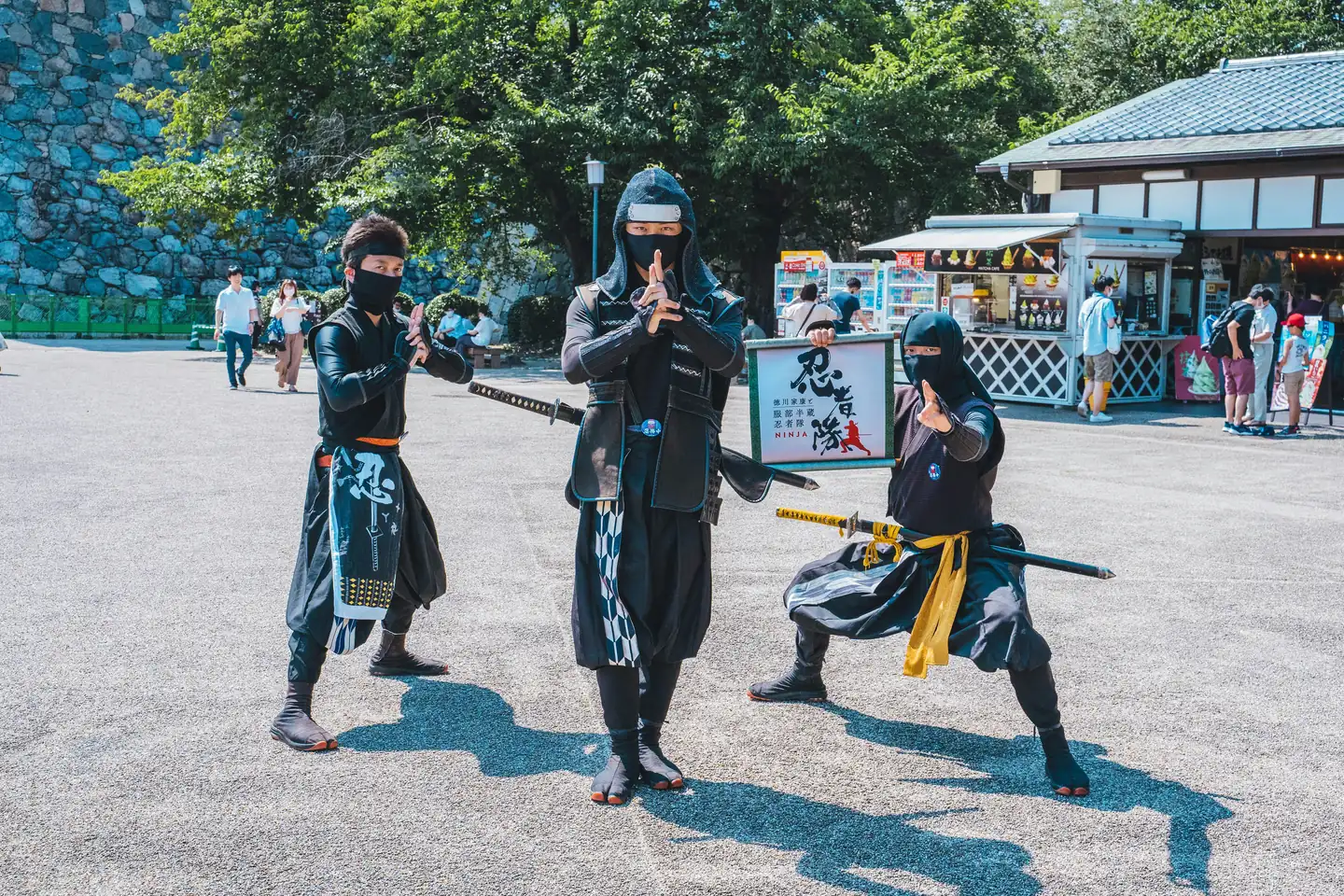 Three ninja performers in black hoods, tabi boots and sashes strike fighting poses with swords at Nagoya Castle, one beside a ninja sign; a ninja troupe in Japan, not the cast of this Shinjuku show