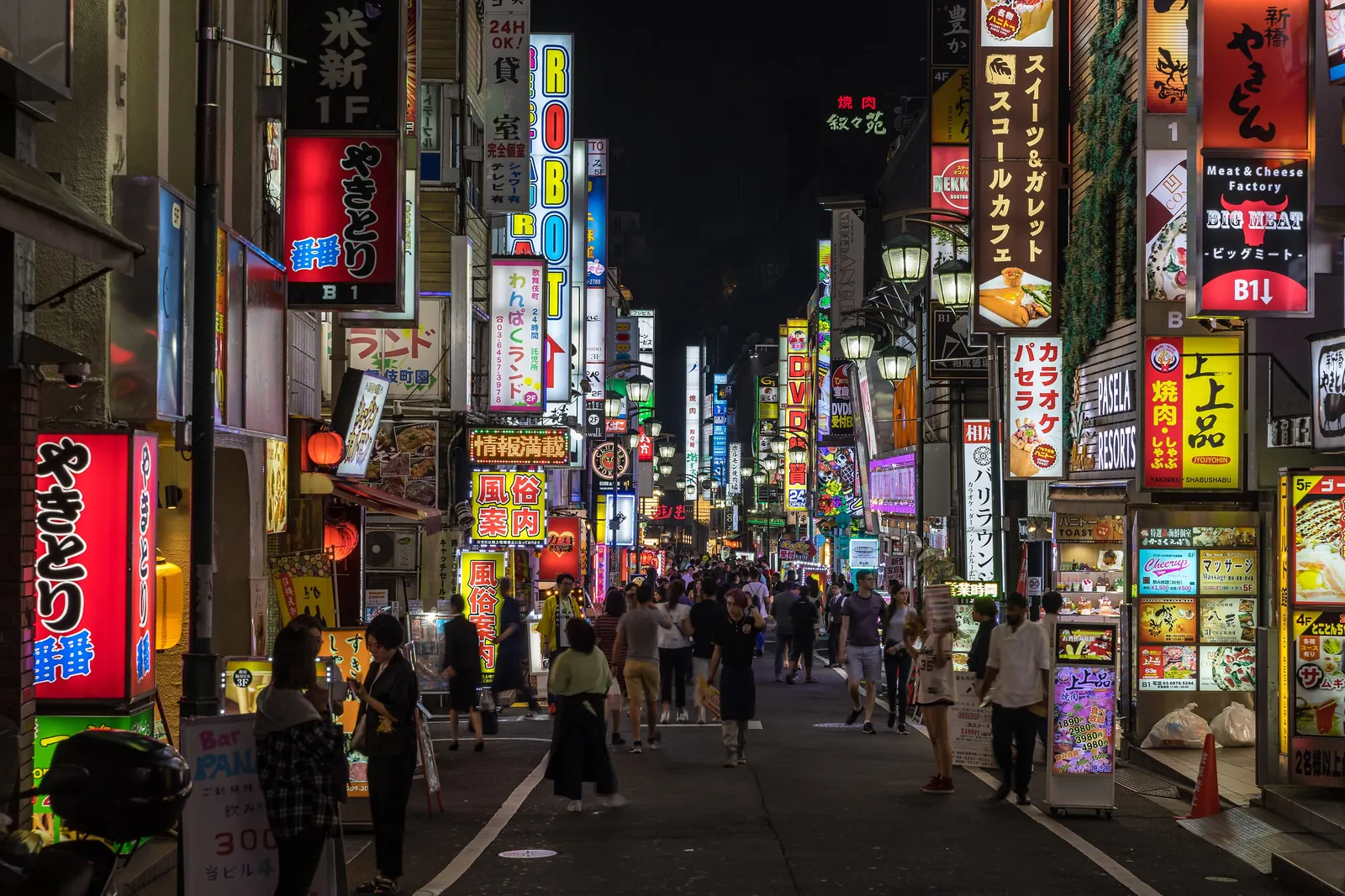 A narrow Kabukichō street at night lined with illuminated vertical signs and crowds in the distance